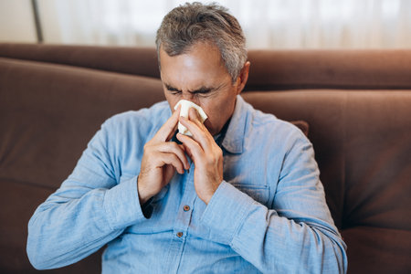 Sick man with runny nose isolated himself at home. Stay home concept. Adult man blows his nose, sits on sofa in living room. Isolate yourself if you get sick. Casual clothes.の写真素材