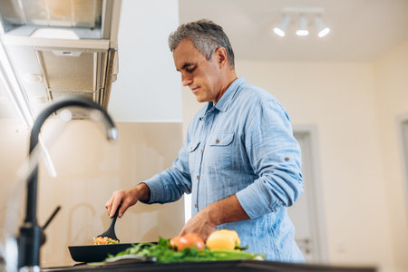 Mature man in blue shirt cooking on modern kitchen. Light cozy room on background. Concentrated man standing by stove, stews vegetables in a frying pan. Healthy food. Bright vegetables on front.の写真素材