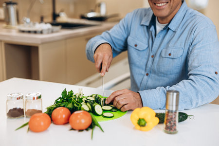 Adult man chops vegetables and greens on a kitchen table. Cut view. Cucumbers, tomatos, peppers, zucchinis and spice jars on clean white table. Cooking with joy concept.の写真素材