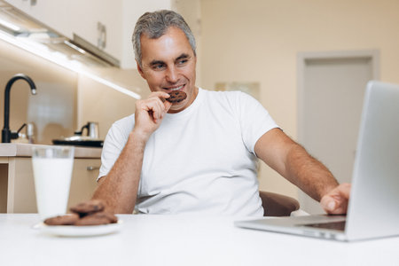 Mature man sitting on kitchen, using his modern gray laptop, watching funny series or videos, laughing and eating yummy cookies. Glass of fresh milk and biscuits on white table.の写真素材