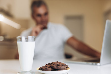 Healthy and tasty breakfast Focus on glass of milk and cookies with chocolate drops. Morning routine concept. Blurred adult man in white t-shirt using laptop and enjoying biscuits.の写真素材