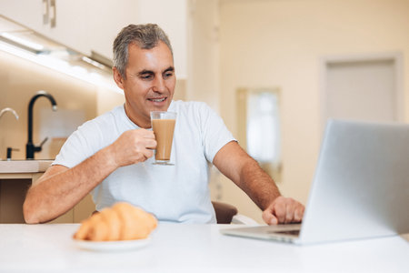 Mature man enjoing his cappuccino and yummy croissant. Morning routine concept. A male freelancer is enjoying his sweet breakfast, looking at laptop monitor and happy that he has found a new customer.の写真素材