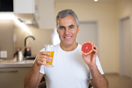 Good loking senior man breakfasts. Vegan food. Mature man in white casual t-shirt holding half of grapefruit and glass of citrus juice and smiling with teeth. Light modern kitchen on background.の写真素材