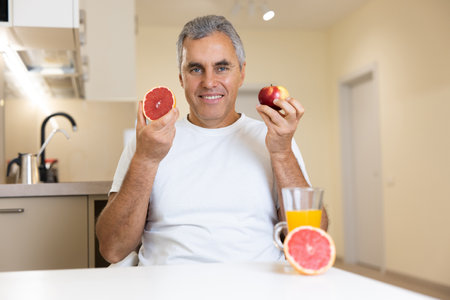 Good looking mature man eats fruits. Organic healthy food concept. Cheerful man holding apple and grapefruit near face and smiling to camera. Glass of fresh orange juice and half of citrus on table.の写真素材