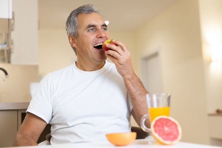 Happy adult man eating fruits and healthy food on breakfast. Morning routine concept. Cut halves of grapefruit and glass of orange juice on table. Cozy modern kitchen on background.の写真素材