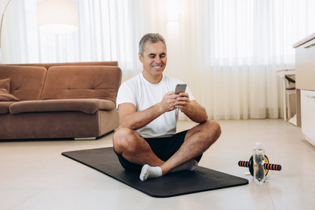 Elderly lazy man laughing when reading memes on smartphone. Put your phone down if you want a strong healthy body Procrastination during fitness. Senior man sitting on yoga mat and texting on phone.の写真素材