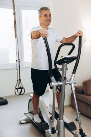 Senior man excercising on cross-trainer and looking away. Healthy and active lifestyle. Workout at home. Cardio fitness using elliptical trainer. White living room on background. Sports equipment.の写真素材