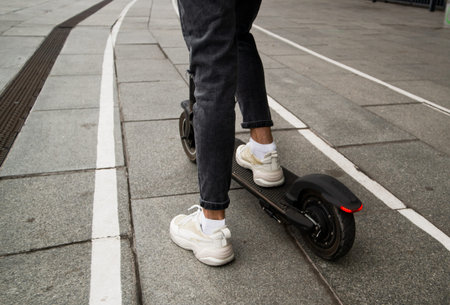 Legs of anonymous guy riding modern electric scooter on asphalt path on blurred background of city...