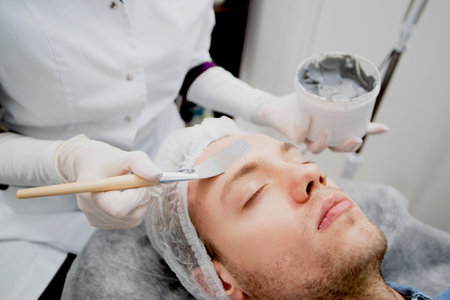 Cosmetologist is putting black mask on the young mans face in the beauty salonの写真素材