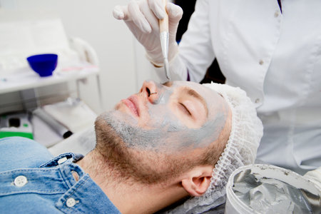 Cosmetologist is putting black mask on the young mans face in the beauty salonの写真素材