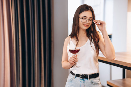 Young and beautiful lady in glasses is drinking red wine at home. Freestyle woman. Freelancer female...