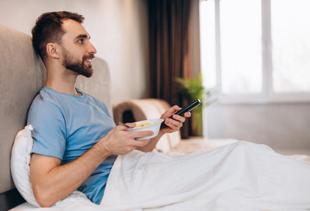 Handsome young man with beard having breakfast in bed and watching tv. weekend lazy day off.の写真素材