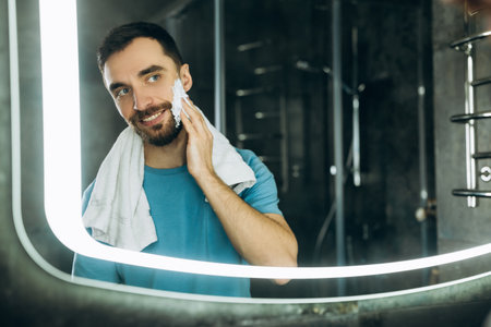 Close-up shot of attractive young man with towel in his neck standing in front of mirror and applying moisturising cream to his face bathroom morning.の写真素材