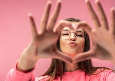 Photo of attractive young woman in glasses shows heart with fingers, blow kiss. Wears casual pink t-shirt white pants isolated pink color backgroundの写真素材
