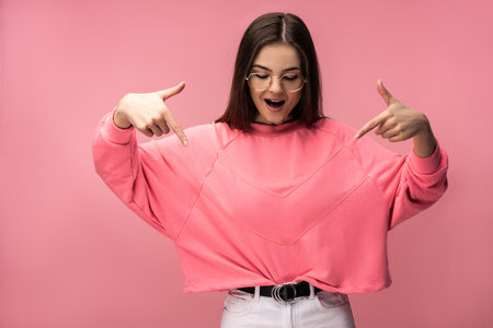 Photo of attractive young woman in glasses points down on something and surprised. Wears casual pink t-shirt white pants isolated pink color backgroundの写真素材