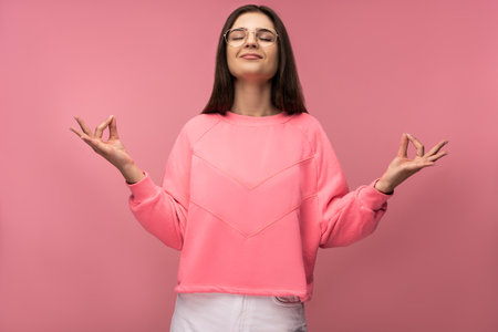 Photo of attractive young woman in glasses, meditation and relax, wears casual pink t-shirt white pants isolated pink color backgroundの写真素材