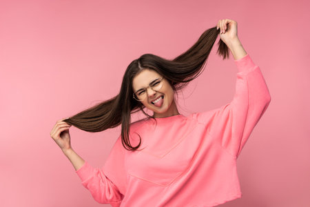 Photo of attractive woman in glasses having fun holds hair wear casual pink t-shirt white pants isolated pink color backgroundの写真素材