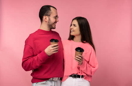Photo of attractive man wih beard in red clothing and woman in pink holds coffee and gawp on each other. Couple looks happy, isolated over pink backgroundの写真素材