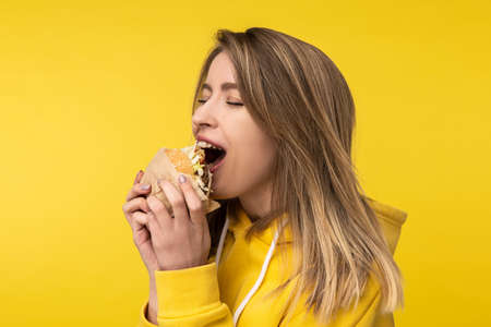 Photo of attractive lady poses in a surprised way bites burger. Wears casual yellow hoody, isolated yellow color backgroundの写真素材