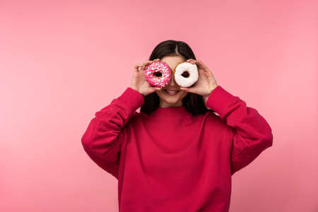 Photo of attractive lady holds covered eyes by donuts candies sweet food wear smile happy casual pink sweater isolated pink color backgroundの写真素材
