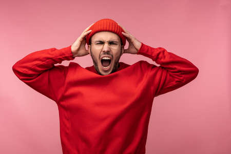 Photo of attractive man with beard in glasses and red clothing. Male screams and yells holds his head, isolated over pink backgroundの写真素材