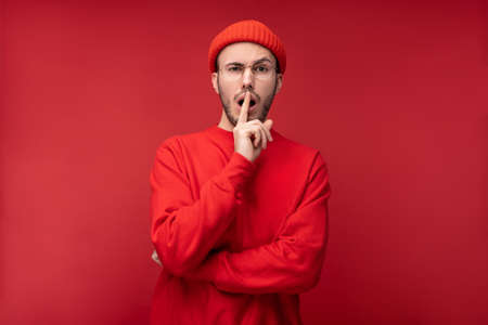 Photo of attractive man with beard in glasses and red clothing. Male shocked and curious, isolated over red backgroundの写真素材