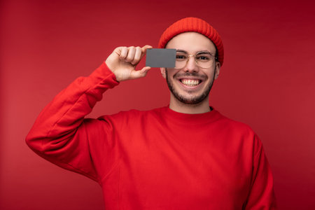 Photo of attractive man with beard in glasses and red clothing. Happy man holds credit card in front of face, isolated over red backgroundの写真素材