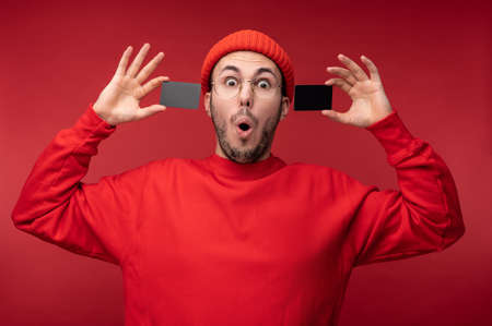 Photo of attractive man with beard in glasses and red clothing. Surprised man holds 2 credit cards, isolated over red backgroundの写真素材