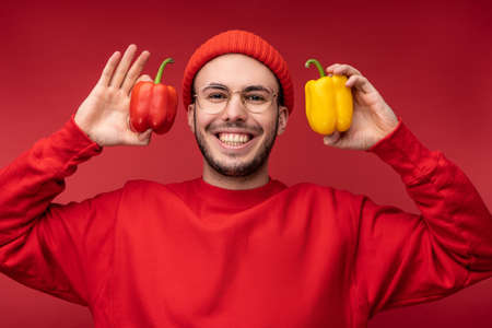 Photo of attractive man with beard in glasses and red clothing. Male holds papers in hands, shows love to vegetables isolated over red backgroundの写真素材