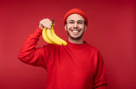 Photo of happy man with beard in glasses and red clothing. Holds bananas and smiles, healthy lifestyle, isolated over red backgroundの写真素材