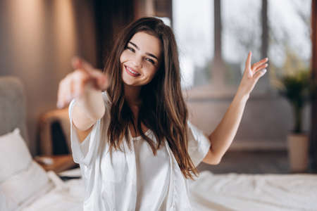 Young beautiful woman in white silk pajamas smiling and looking at the camera. Beautiful female model in a cheerful mood is dancing and smilingの写真素材