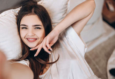 Young beautiful woman smiles while taking a selfie and holds her hand near her face. The brunette makes a selfie in the bed with a smileの写真素材