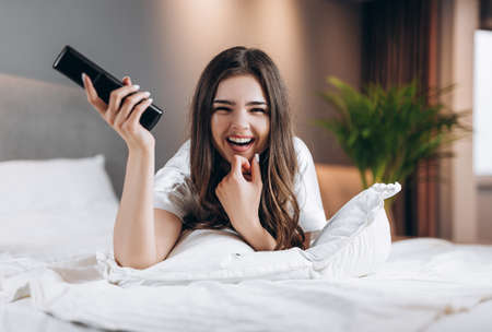 woman in white silk pajamas watches TV in the bedroom on the bed. Female model with a tv remote control in her hands lying on the bed. morning or evening in the bedroom with a female modelの写真素材