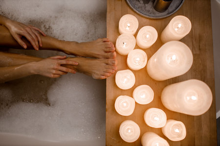 Close up photo of womans body in spa bathtub with foam and wooden table vase of plant candle. Modern...