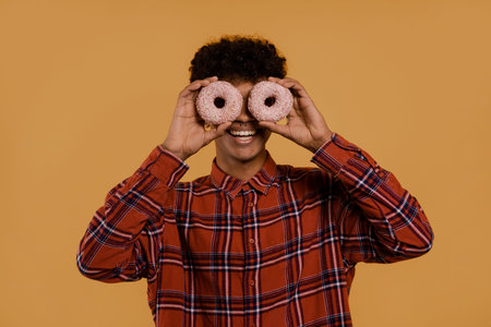 Photo of cute African male farmer looks happy and covers his eyes up by donuts. Man wears plaid...