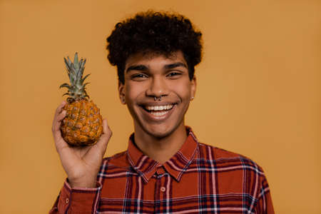 Photo of cute African male farmer holds pineapple and slime. Man wears plaid shirt, isolated brown color backgroundの写真素材