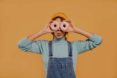 Close up photo of cute female farmer covers her eyes by donuts. Wears denim overalls and hat, isolated brown color backgroundの写真素材
