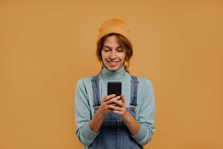 Photo of cute female farmer holds smartphone and seems happy. Wears denim overalls and hat, isolated brown color backgroundの写真素材