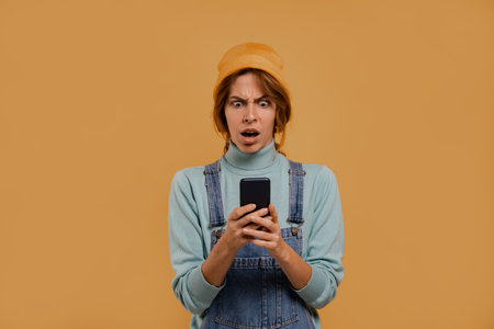 Photo of cute female farmer holds smartphone and looks like something goes wrong. Wears denim overalls and hat, isolated brown color backgroundの写真素材