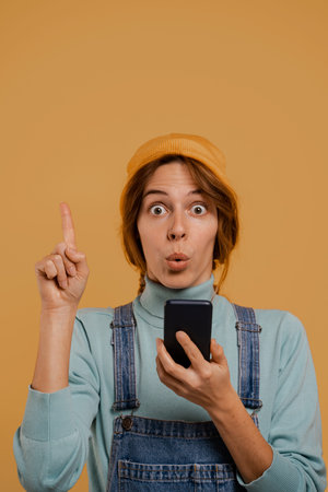 Photo of cute female farmer holds smartphone and surprised with idea she has got. Wears denim...