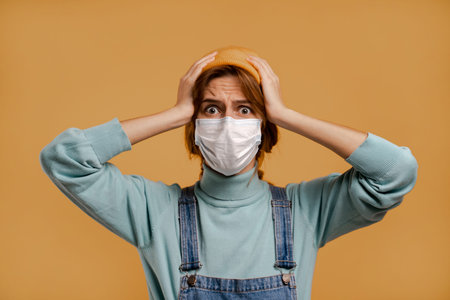 Photo of cute female farmer holds head with disappointment face mask for quarantine time. Wears...