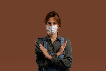 Photo of cute female farmer with face mask shows lockdown and quarantine time. Wears denim overalls,...