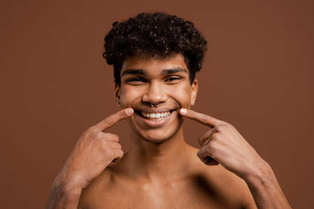 Photo of attractive black man with piercing show how wide his smile is. Naked torso, isolated brown color backgroundの写真素材