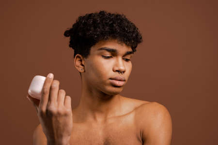 Photo of attractive black man with piercing holds nutrition cream and looks down. Naked torso, isolated brown color backgroundの写真素材