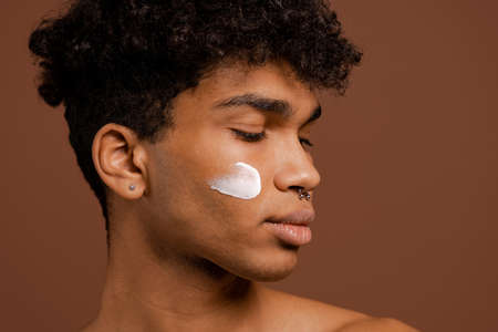 Photo of attractive black man with piercing and nutrition cream on face. Naked torso, isolated brown color backgroundの写真素材