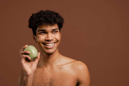 Photo of attractive black man with piercing holds apple and smiles. Naked torso, isolated brown color backgroundの写真素材