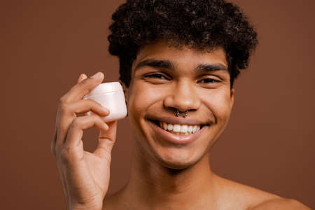 Close up photo of attractive black man with piercing holds nutrition cream for face. Naked torso, isolated brown color backgroundの写真素材