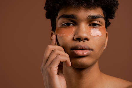 Close up photo of attractive black man with piercing looks at camera and touches his face with eye patches on. Naked torso, isolated brown color backgroundの写真素材