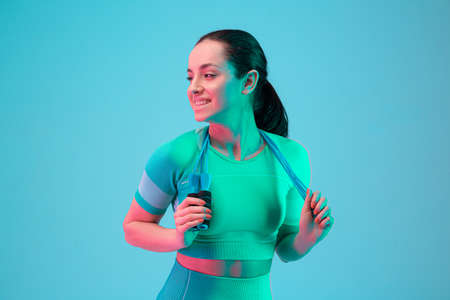Young sportive woman training with skipping rope isolated on gradient studio background in neon light. Modern sport, action, motion, youth concept. Beautiful caucasian woman practicingの写真素材