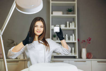 Consultation in cosmetology clinic. Female beauty doctor in white coat and black gloves shows thumb up on cream. Healthcare concept.の写真素材